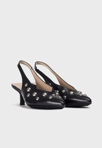 Zapatos de slingback de piel negra con punta afilada, tacón pequeño y tachuelas plateadas decorativas en la parte frontal. Textura suave, diseño minimalista.