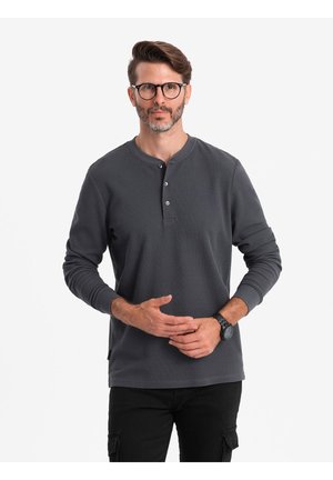 Chemise henley à manches longues gris foncé avec trois boutons à l'avant et des poignets côtelés, portée avec un pantalon noir. Texture lisse et design à coupe décontractée.