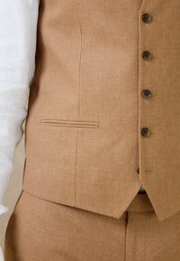 Gros plan sur une personne portant un gilet de costume beige à quatre boutons sur une chemise blanche, montrant la texture du tissu et le détail de la poche sur mesure.