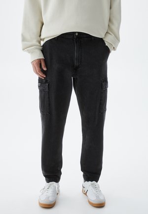 Personne portant un pantalon cargo noir avec des poches latérales, un sweat-shirt blanc et des baskets blanches, debout devant un fond uni clair.