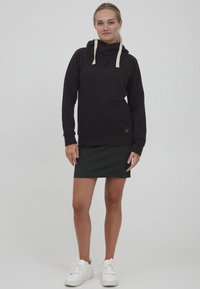 Sudadera con capucha negra y cordones crema, con bolsillo estilo canguro y un parche de logo. Llevada sobre una falda de punto verde y zapatillas blancas.