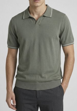 Uomo che indossa una polo a maglia a maniche corte verde oliva con rifiniture bianche su colletto e maniche, abbinata a pantaloni grigio scuro.