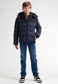 Geacă matlasată bleumarin cu fermoar frontal și glugă, cu modele matlasate orizontale. Purtați cu blugi albaștri și adidași alb-negru.