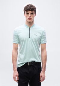 Νεαρός  άνδρας που φοράει κοντομάνικο πόλο σε ανοιχτό μέντα πράσινο χρώμα με μαύρο φερμουάρ και λογότυπο "BOSS", συνδυασμένο με μαύρο παντελόνι και μαύρη ζώνη.