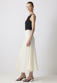 Zwarte mouwloze top gecombineerd met een witte, vloeiende rok. De rok heeft een gladde textuur; accessoires bestaan uit gouden creolen en zwarte sandalen.