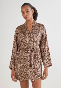 Leopardmönstrad rock i satinmaterial, med ett knytbälte, vida ärmar och sjalkrage. Färgskalan inkluderar nyanser av brunt och svart.