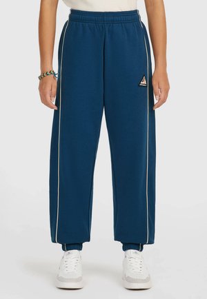 Person wearing blue elastic-waist sweatpants with white side stripes and elastic cuffs, paired with white sneakers and a woven bracelet.