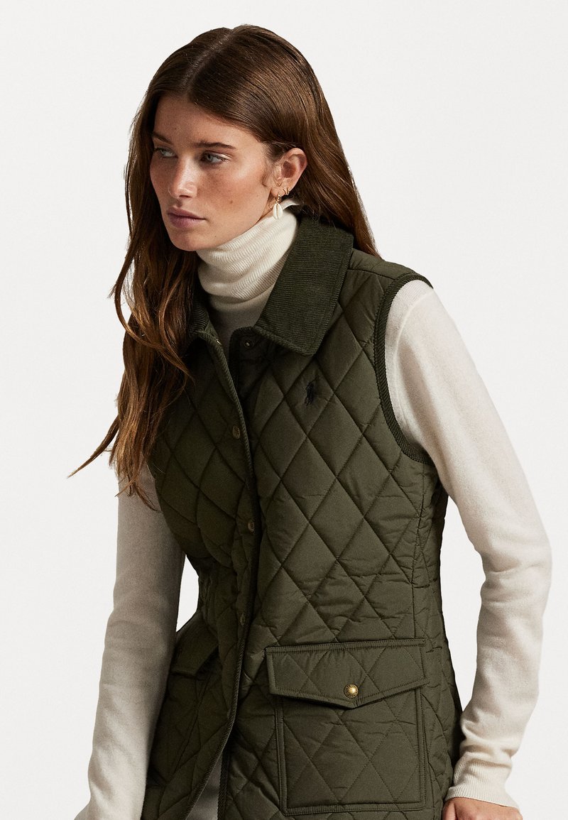 Grünes Steppweste mit einem Cordkragen, ausgestattet mit einer Vordertasche und goldenen Druckknöpfen, über einem cremefarbenen Rollkragenpullover getragen.