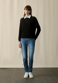 Nainen seisoo beige taustalla mustassa hupparissa, jossa on valkoinen kaulus, sinisissä farkuissa ja valkoisissa tennareissa, kädet rentoina sivuilla.