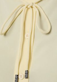 Jasnożółta bluzka z zapięciem na supeł, ozdobiona guzikiem i teksturowaną tkaniną, podkreślona czarno-białymi paskowanymi dodatkami.