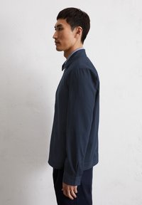 Navy blue lightweight jacket with a front pocket, folded collar, and long sleeves, shown on a male model in profile against a plain background.