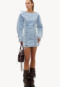 Femme portant une robe en denim bleu clair avec des détails à lacets, tenant un sac à main marron foncé et portant des bottes marron foncé décontractées.