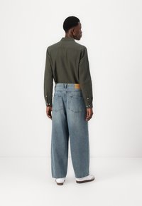 Mörkgrön långärmad skjorta som kombineras med ljusblå vida jeans och vita skor. Synlig sömnad och fickor på jeansen.