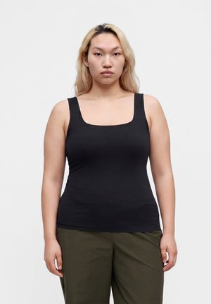 Ung person med langt blondt hår iført en sort tanktop og olivengrønne bukser, der står mod en ensfarvet hvid baggrund.