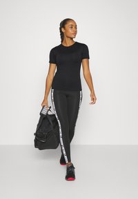 Svart tettsittende t-skjorte kombinert med svarte leggings med hvite sidestriper. Modellen bærer en svart treningsveske og har på seg svarte joggesko.