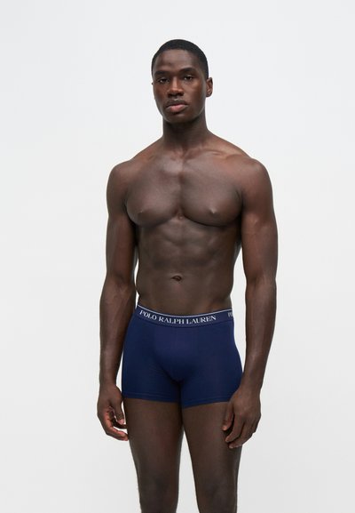 Μυώδης άνδρας που φοράει σκούρα μπλε boxer briefs Polo Ralph Lauren, στέκεται μπροστά σε απλό λευκό φόντο.