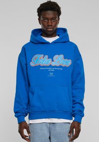 Sininen huppari, jossa etutasku, jossa on punaiset ja valkoiset "Fake Love" -tekstit sekä "Upscale Studios 143 Collection" -logo. Puuvillakangasta, rento malli.