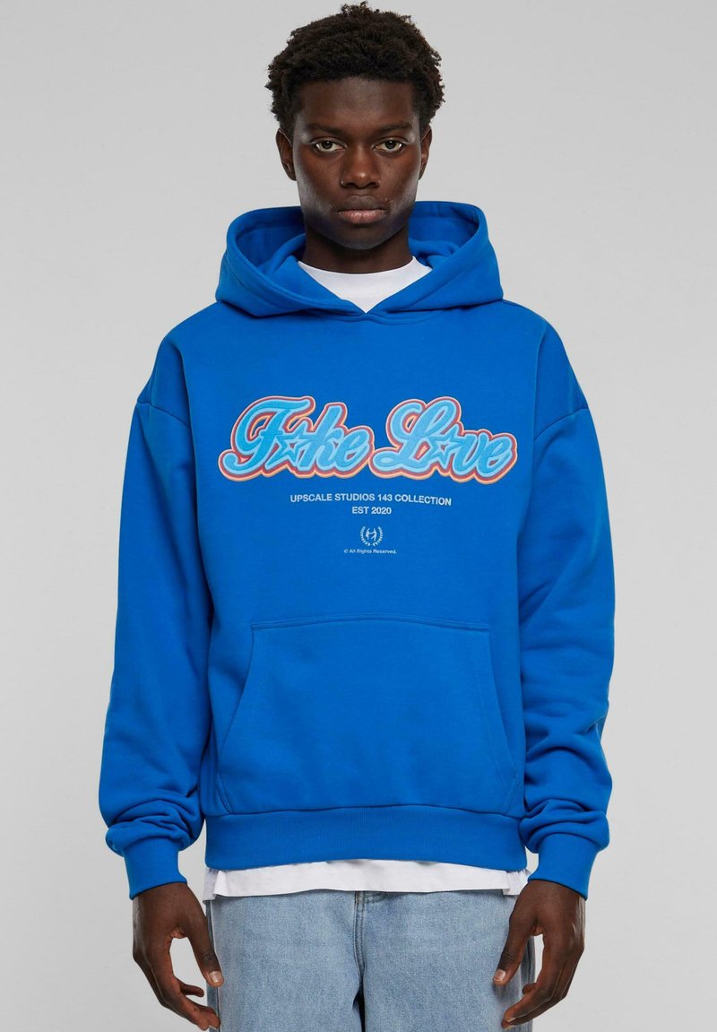 Sininen huppari, jossa etutasku, jossa on punaiset ja valkoiset "Fake Love" -tekstit sekä "Upscale Studios 143 Collection" -logo. Puuvillakangasta, rento malli.