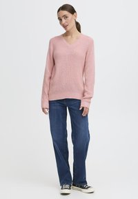 Maglione rosa lavorato a maglia con scollo a V, polsini e orlo a coste. Abbinato a jeans blu a gamba dritta e scarpe da ginnastica nere.