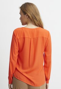 Långärmad blus i stark orange, tillverkad av mjukt tyg. Har en rundad halsringning och en något böjd nederdel med detaljer på bakfaltet.