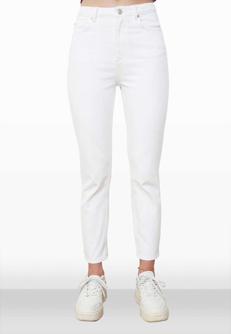 Personne portant un jean skinny blanc taille haute, associé à des baskets blanches et un bracelet de cheville doré à la cheville gauche, debout sur un fond clair.