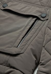 Quilted brown jacket with a textured surface, featuring a large front pocket with a flap and a zippered side pocket.
