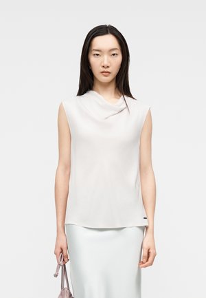 Donna con lunghi capelli scuri che indossa una canotta bianca senza maniche e una gonna, che tiene una piccola borsa rosa chiaro su uno sfondo semplice.