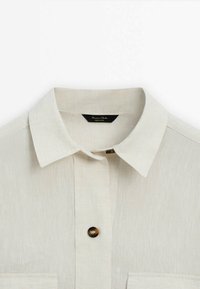 Beige linen shirt with a classic collar, front button closure, and a chest pocket. Features a textured fabric with a smooth finish.