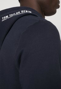 Granatowa bluza z kapturem z teksturowanej tkaniny, okrągłym dekoltem i białym napisem marki z tyłu, podkreślającym nazwę marki i szczegóły.
