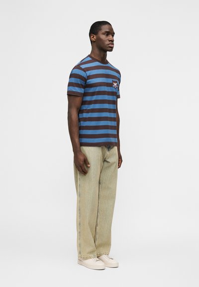 Άνδρας που στέκεται φορώντας μπλε και κ αφέ ριγέ κοντομάνικη μπλούζα, ανοιχτό μπεζ τζιν με φαρδιά πόδια και λευκά αθλητικά παπούτσια σε λευκό φόντο.