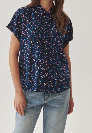 Femme portant une chemise boutonnée bleu marine avec des motifs de feuilles roses et bleues, et un jean en denim bleu clair, debout devant un fond uni.