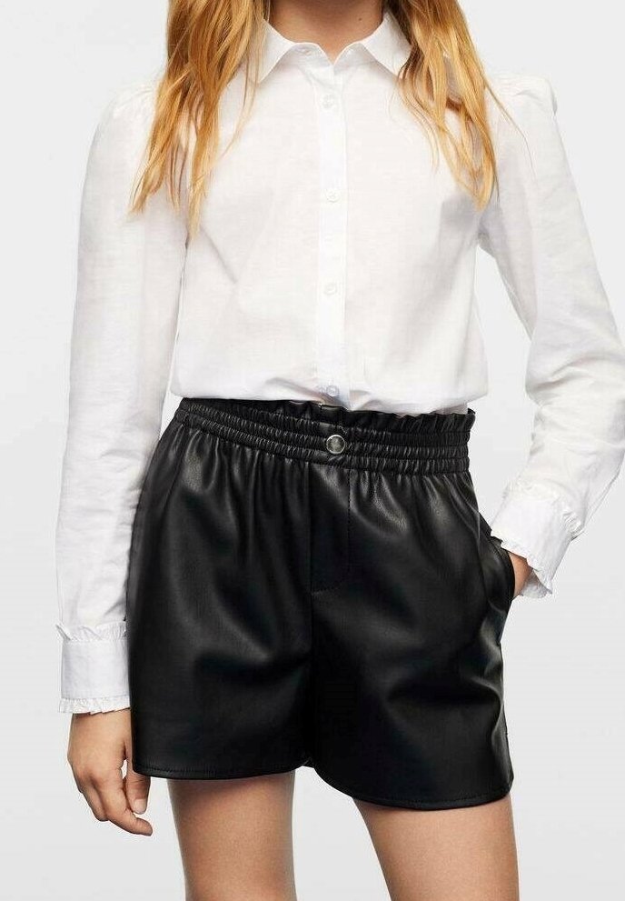 Femme portant une chemise blanche à boutons rentrée dans un short en cuir noir taille haute avec une ceinture élastique et des poches latérales.