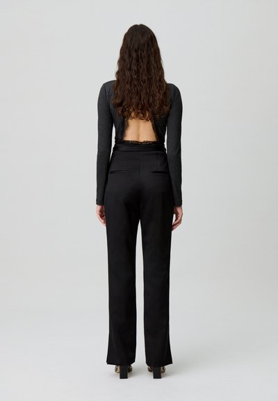 Donna con lunghi capelli ricci di spalle, che indossa pantaloni neri a vita alta e un top nero a maniche lunghe con schiena scoperta e bordi di pizzo.