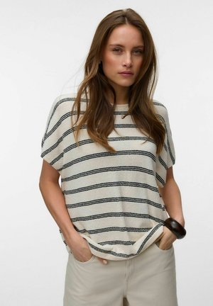 Femme aux longs cheveux bruns portant un haut crème à manches courtes avec des rayures horizontales noires et un pantalon beige, mains dans les poches, sur fond blanc.