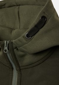 Olivgrön ziphoodie med texturerad tyg, som har dragsko och svarta detaljer på kragen och dragkedjan. Slät ytdetalj.
