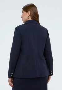 Frau mit langen braunen Haaren, die ein dunkelblaues Blazer und einen passenden Rock trägt, von hinten vor einem schlichten weißen Hintergrund gezeigt.