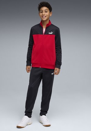 Niño de pie sonriendo, vestido con un chándal Puma rojo y negro y zapatillas blancas, sobre un fondo gris liso.