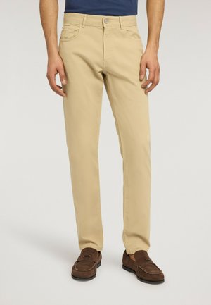 Mann trägt beigefarbene, gerade geschnittene Hose mit Seitentaschen, Knopf- und Reißverschluss, kombiniert mit braunen Wildleder-Loafers und einem marineblauen Hemd.