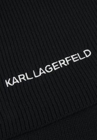 Tecido de malha ribbed preto com texto bordado em branco "KARL LAGERFELD." A textura apresenta linhas verticais proeminentes com um acabamento suave.