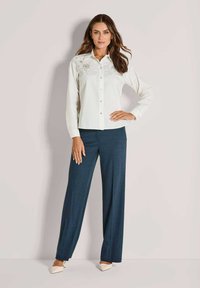 Een vrouw in een witte geborduurde blouse met lange mouwen en donkere wijde broek staat met één hand op haar heup tegen een effen muur.