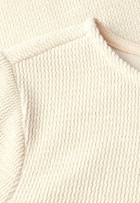 Off-white texturiertes Gewebe mit einem gerippten Muster und einem glatten Ausschnitt. Sichtbare Nahtdetails verleihen dem Gesamtdesign Dimension.