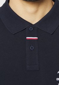 Marinblå polotröja med texturerad tyg, två svarta knappar och en dekorativ rand i rött, vitt och blått på kragen. Broderad logotypdetalj.