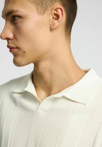 Polo shirt bianco a maglia con un motivo testurizzato, caratterizzato da un colletto classico e un'apertura a placket. Orecchino a cerchio piccolo in argento visibile.