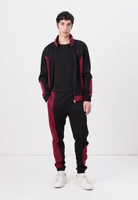 Zwarte en bordeauxrode trainingspak gemaakt van soepele stof; jack met rits, hoge kraag en broek met zijstrepen; gedragen met witte sneakers.