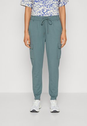 Pantalones cargo verde azulado con cintura ajustable, que cuentan con bolsillos laterales y perneras ajustadas, combinados con zapatillas blancas y una blusa con patrón.
