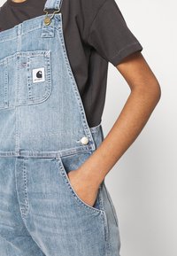 Lichtblauwe denim salopettes met een voorzak, verstelbare banden en een witte logo patch. De stof heeft een lichte textuur met zichtbare stiksels.