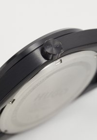 Μαύρο ρολόι με κομψό μεταλλικό σώμα, που διαθέτει υφή στο κορώνα με την επιγραφή "HUGO" και μια ομαλή, ανακλαστική ασημένια πλάτη.