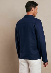 Granatowa lniana koszula z długim rękawem, standardowym kołnierzykiem i mankietami na guziki. Posiada prostą, gładką fakturę oraz dobrze dopasowany krój.
