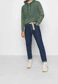 Sweat à capuche vert avec poche avant et cordons, associé à un jean denim bleu et des baskets beige avec des accents blancs.