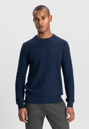 Navy blue knitted sweater featuring a crew neckline, long sleeves, ribbed cuffs, and a subtle texture. Paired with gray pants.